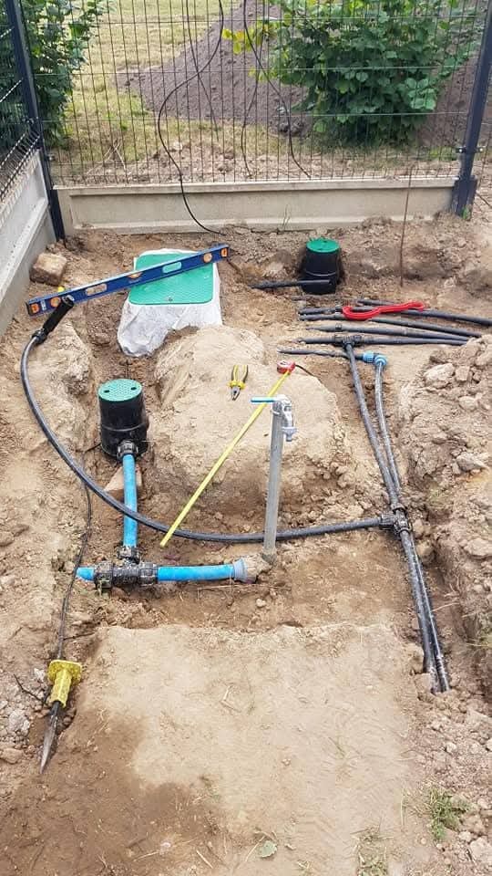 Instalacja systemu nawadniania w ziemi: rury, złączki, zawór, skrzynka sterująca, poziomica. Widoczne narzędzia i elementy systemu przed zasypaniem.