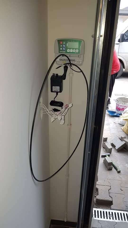 Panel sterowania systemem nawadniania zamontowany na ścianie, widoczne okablowanie i zasilacz. Na podłodze kostka brukowa i narzędzia.