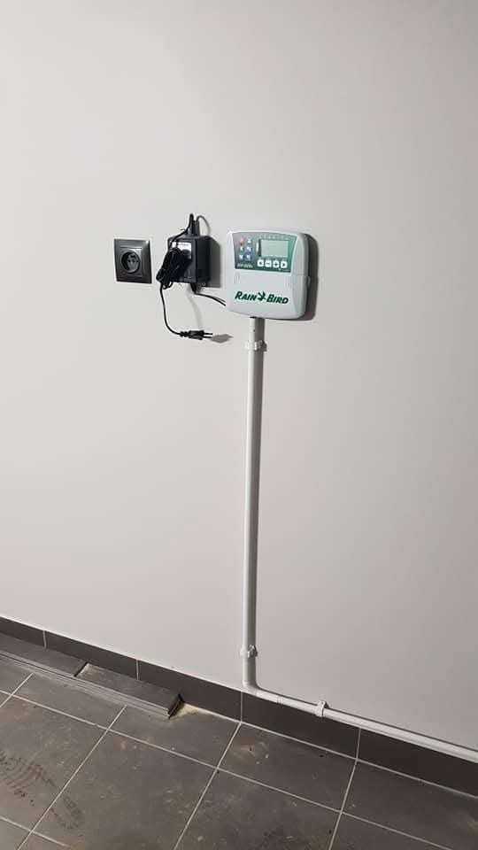 Panel sterowania systemem nawadniania Rain Bird zamontowany na białej ścianie, z widoczną instalacją elektryczną i rurami doprowadzającymi wodę. Szare płytki podłogowe.