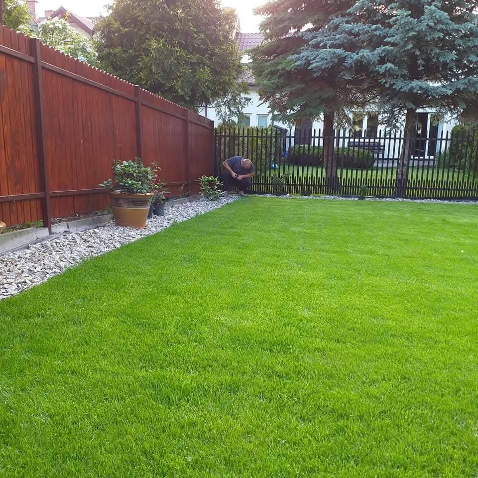 Soczysta, zielona trawa w ogrodzie z drewnianym i metalowym ogrodzeniem. Osoba dogląda roślin przy granicy trawnika, obok donicy z kwiatami.