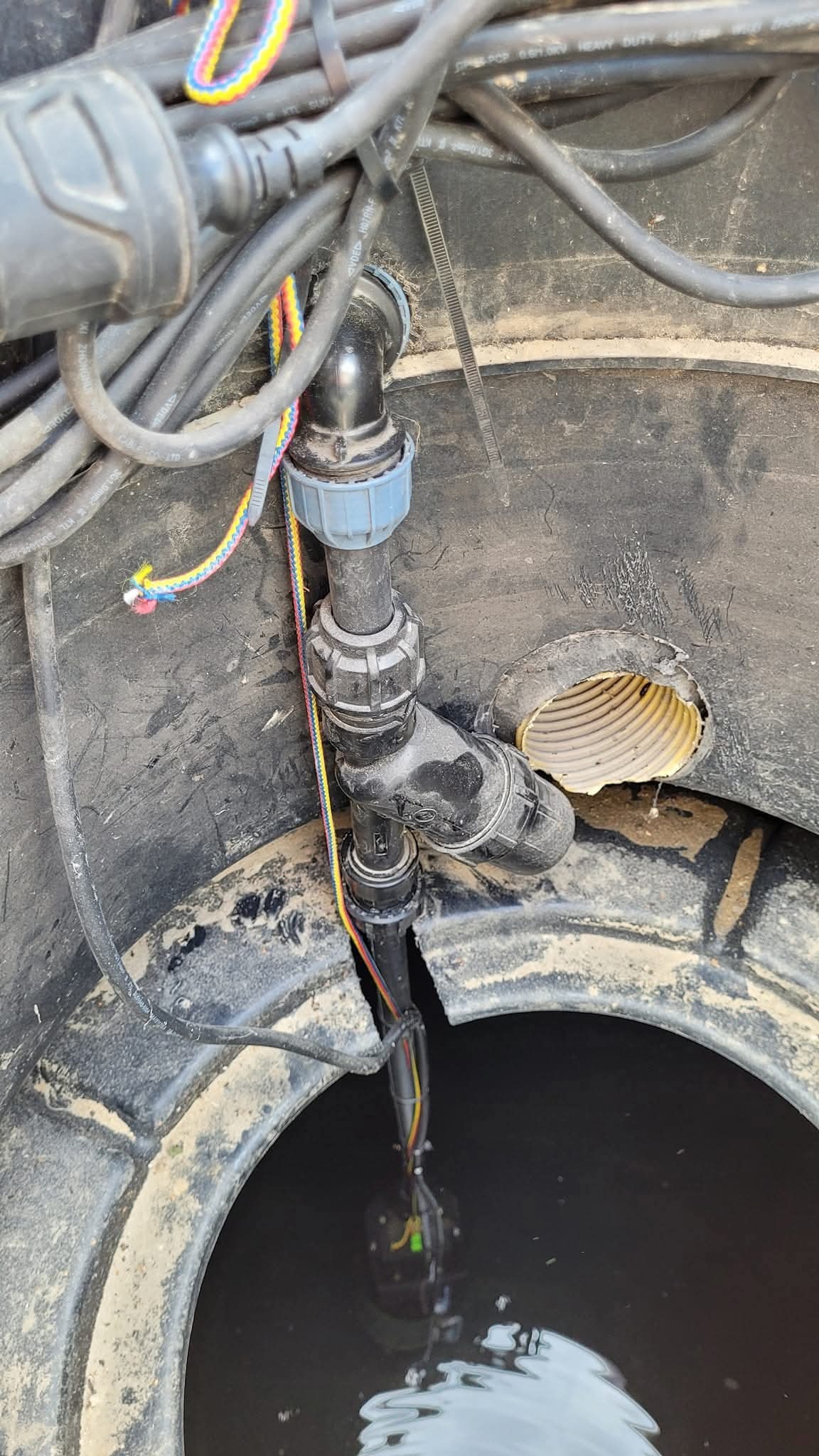 Studnia z pompą zanurzoną w wodzie, widoczne rury i przewody. Instalacja systemu nawadniania w betonowej studni z wywierconym otworem na rurę odpływową.