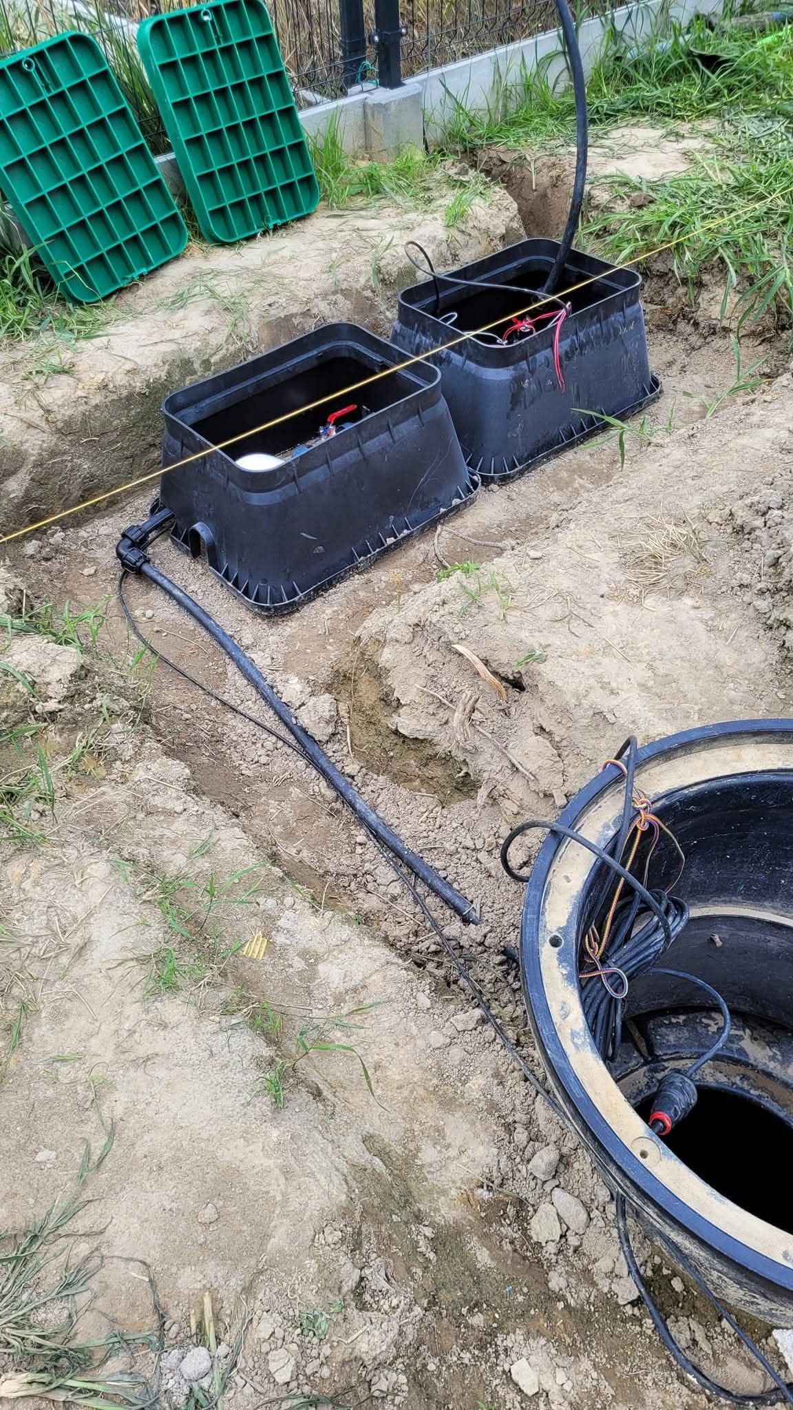 Montaż podziemnego systemu nawadniania: czarne skrzynki zaworowe i rury w wykopie, zielone pokrywy obok. Widoczne okablowanie i elementy instalacji.