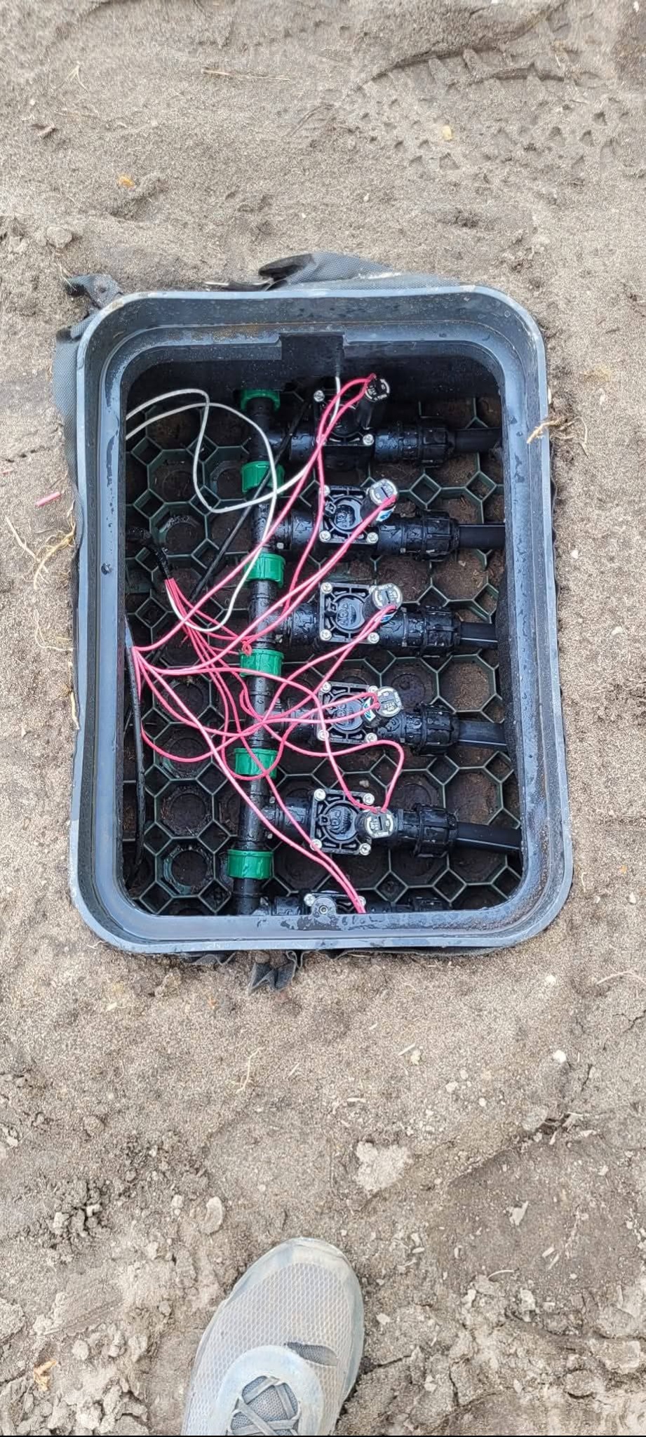 Skrzynka zaworowa z elementami systemu nawadniania, widoczne rury, zawory i przewody elektryczne. Tło stanowi ziemia.