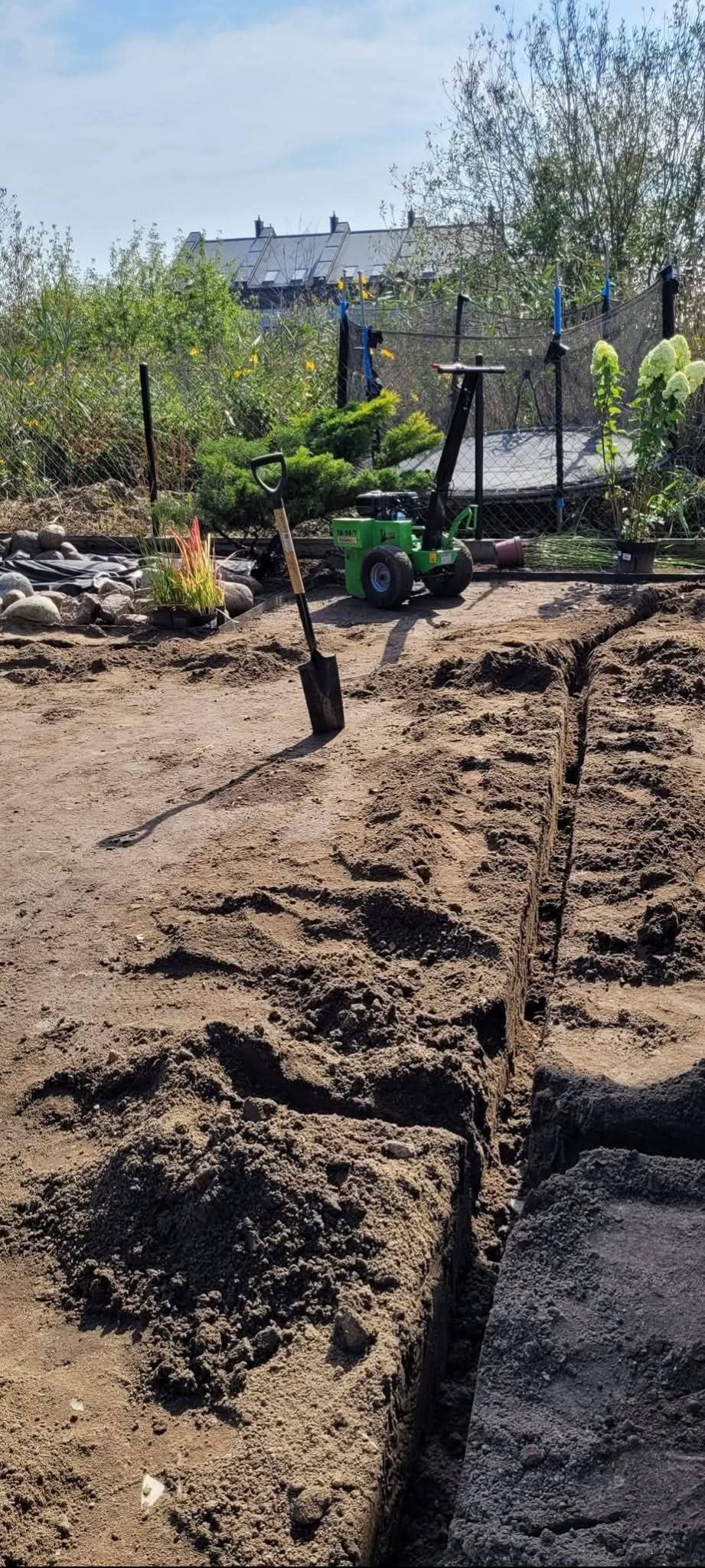 Wykopany rów w ziemi, łopata wbita obok, w tle zielona mini-koparka, rośliny i ogrodzenie. Przygotowanie terenu pod instalację w ogrodzie.