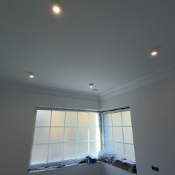 KONOTOP-BUD VIKTOR KONOTOPCHYK - Białe wnętrze w trakcie prac wykończeniowych: okno zabezpieczone folią, widoczne listwy dekoracyjne pod sufitem i oświetlenie punktowe. Ściany przygotowane do malowania.