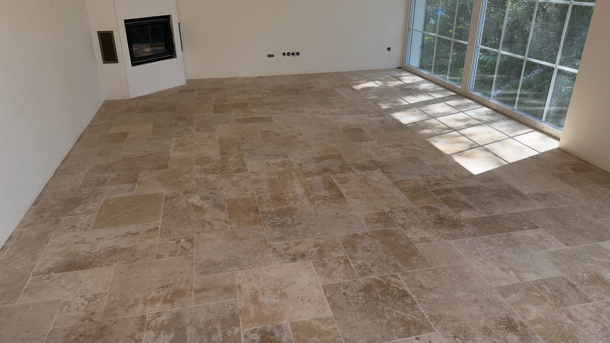 Beżowa, kamienna podłoga ułożona z płytek o zróżnicowanych rozmiarach w jasnym pokoju z dużym oknem i kominkiem. Widoczne refleksy słoneczne na podłodze.