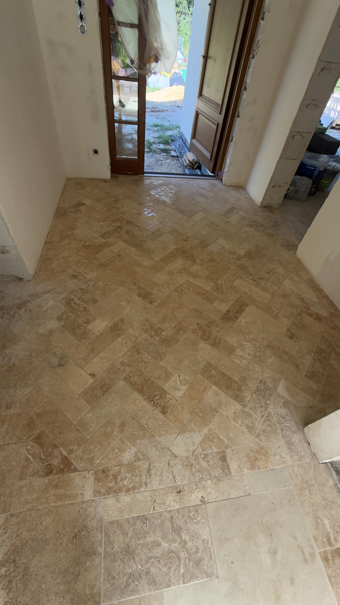 Beżowa podłoga z płytek ułożonych w jodełkę, widok z góry, z otwartymi drzwiami w tle. Częściowo wykończone ściany. Widoczne ślady wilgoci na podłodze.