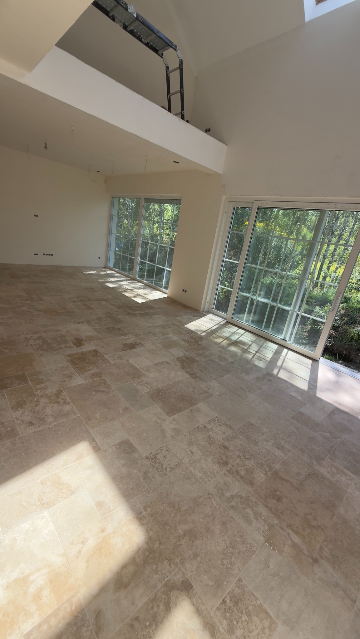 Jasna, przestronna sala z ułożoną podłogą z płytek w odcieniach beżu i brązu. Duże okna wpuszczają naturalne światło. Widoczna drabina w górnej części pomieszczenia.