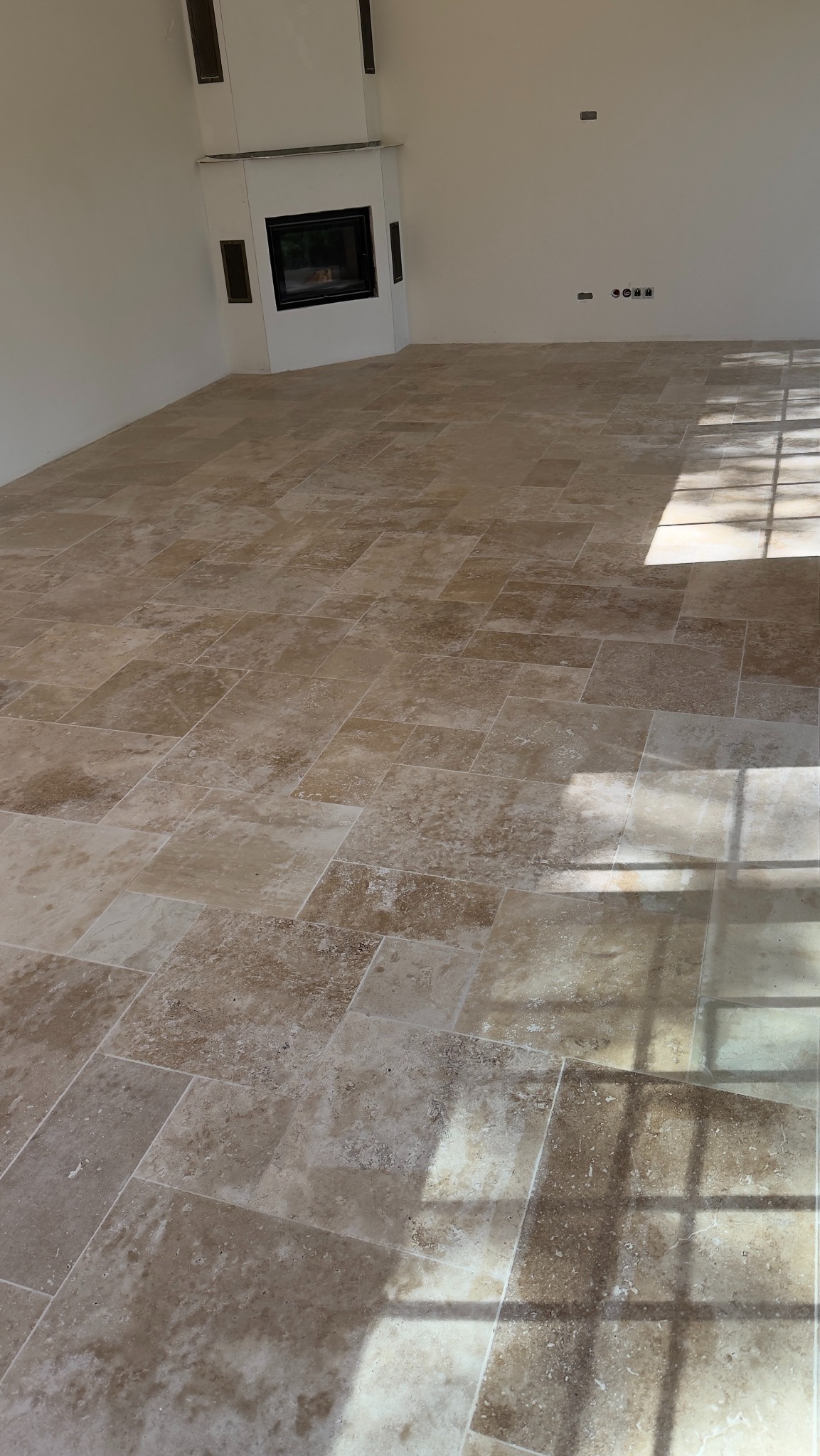 Beżowa podłoga z trawertynu ułożona w salonie z kominkiem. Na podłodze cień okienny. Widoczne gniazdka elektryczne na ścianie.