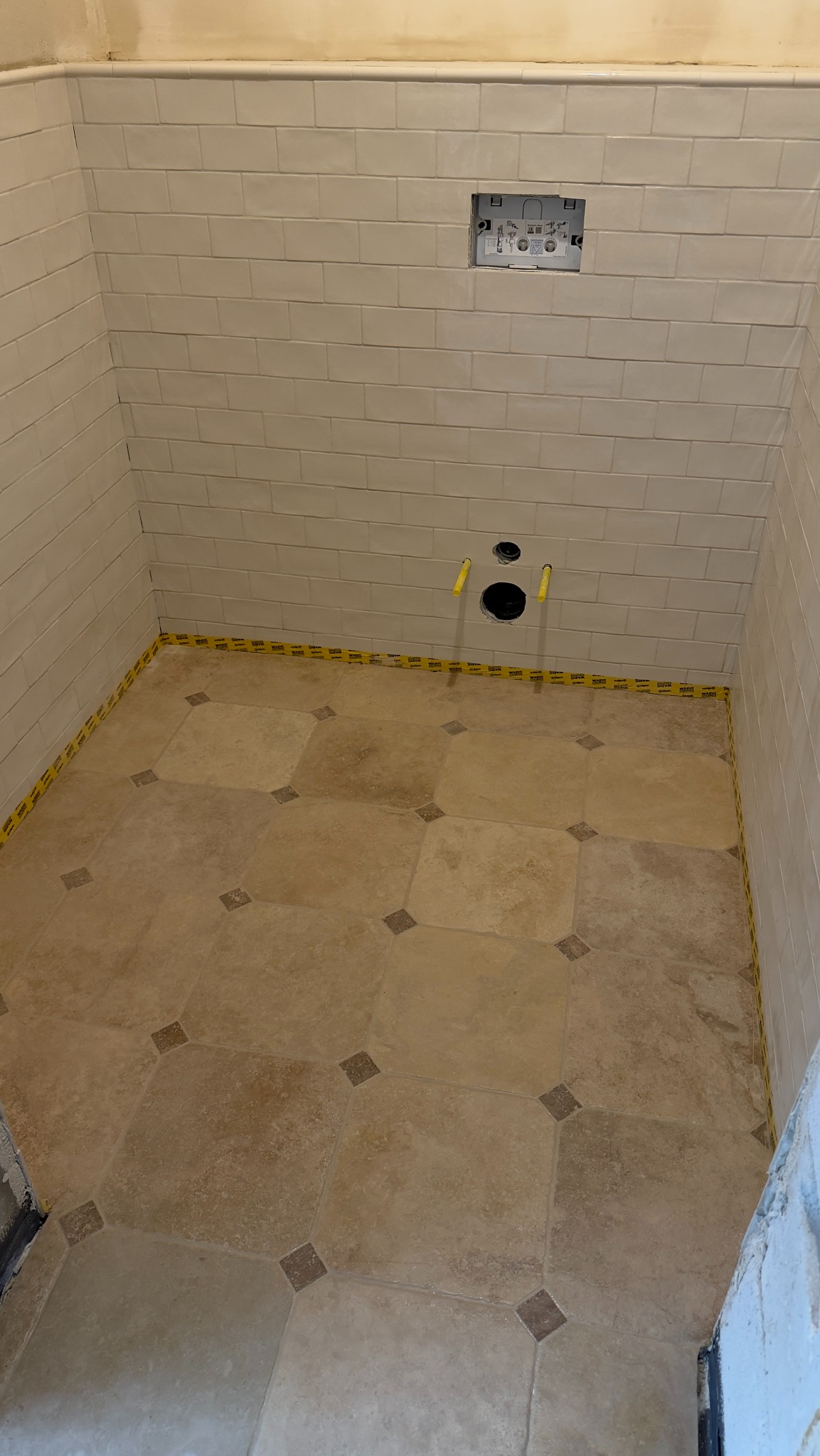 Wykończona łazienka z beżowymi płytkami na ścianach i podłodze w romby. Widoczne otwory na instalacje i taśma zabezpieczająca na styku ścian i podłogi.