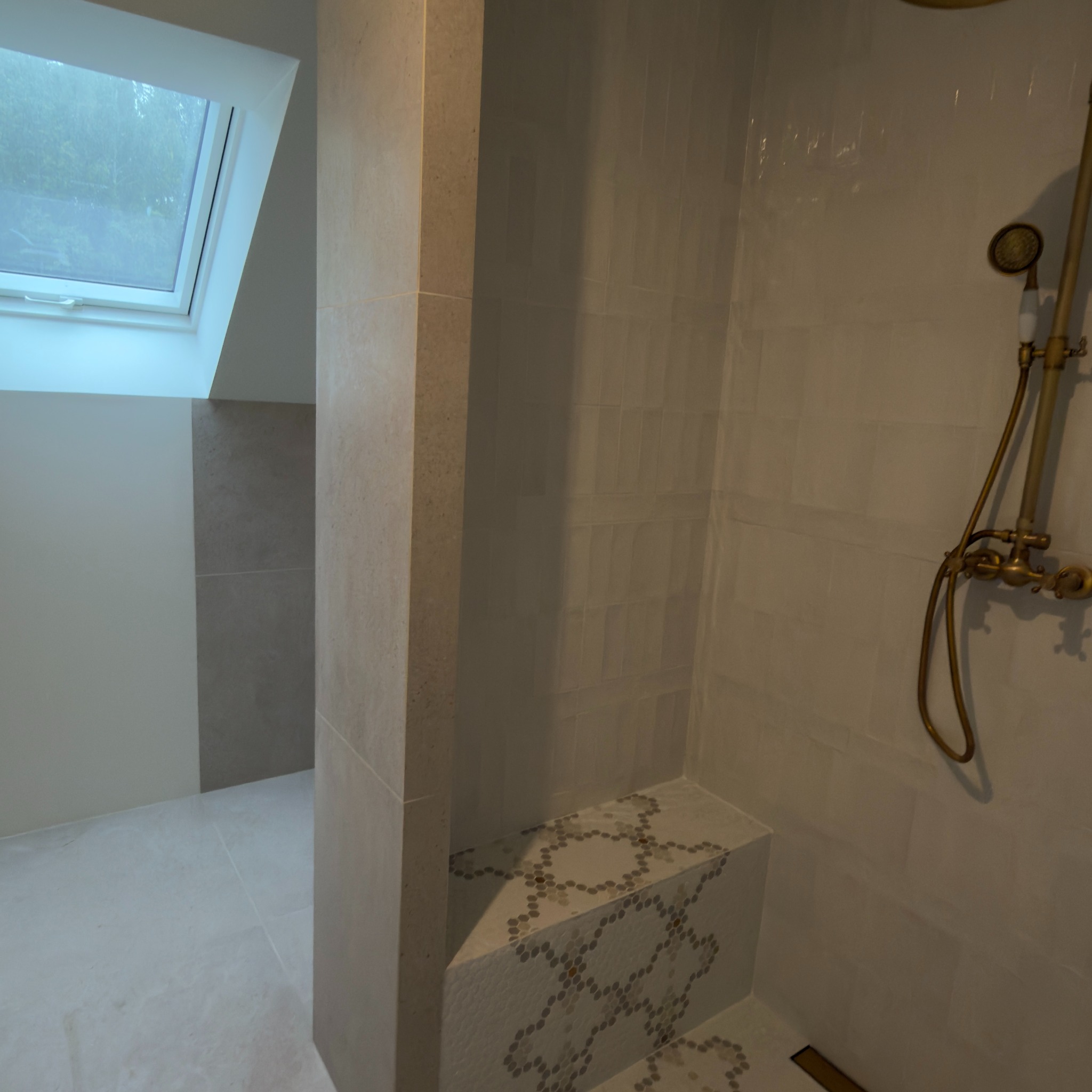 Jasna łazienka z oknem dachowym i kabiną prysznicową wykończoną beżowymi płytkami. Widoczna bateria prysznicowa w stylu retro i mozaika na siedzisku.