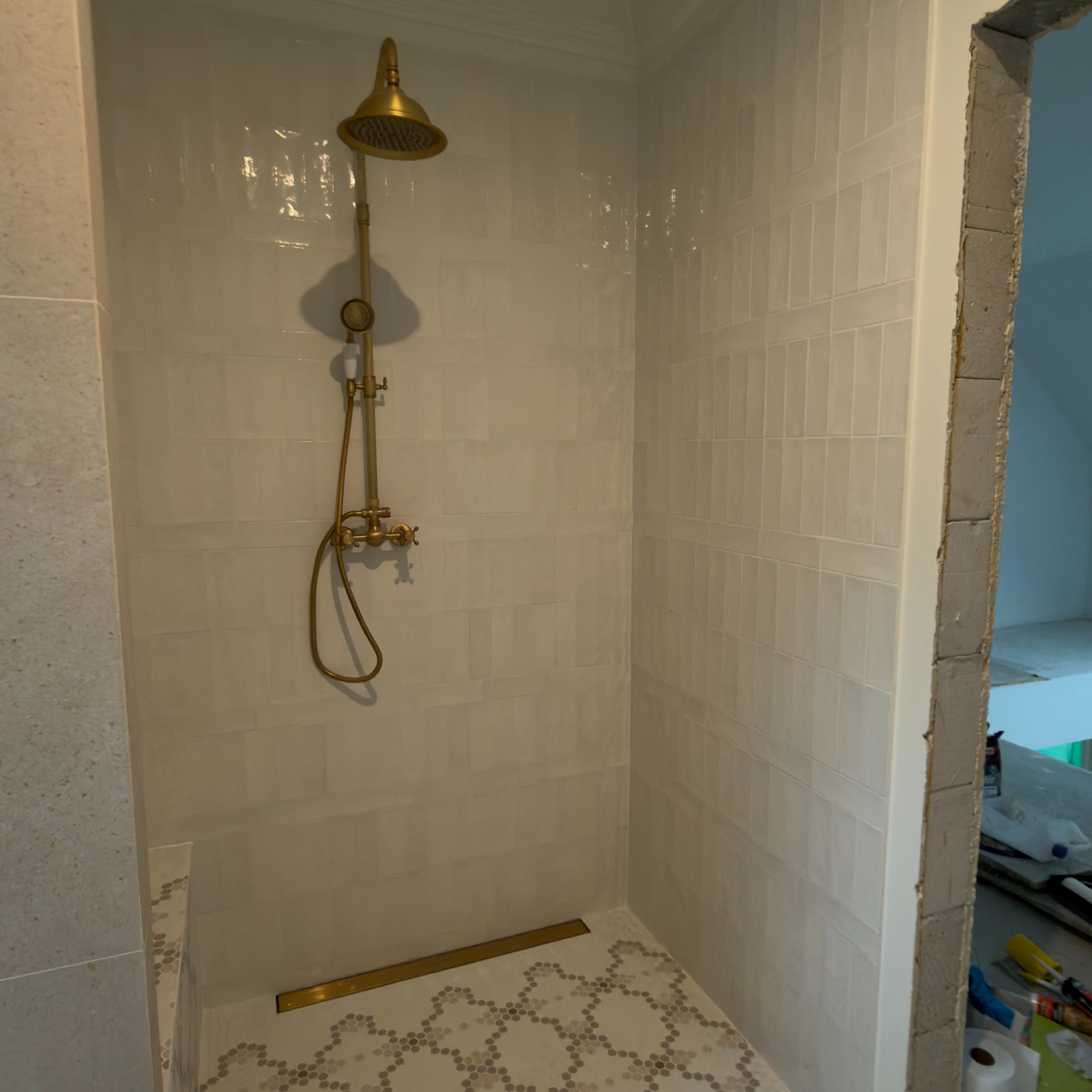 Wykończona kabina prysznicowa z białą, prostokątną glazurą i złotą armaturą. Na podłodze mozaika w odcieniach szarości. Widoczny fragment surowego muru.