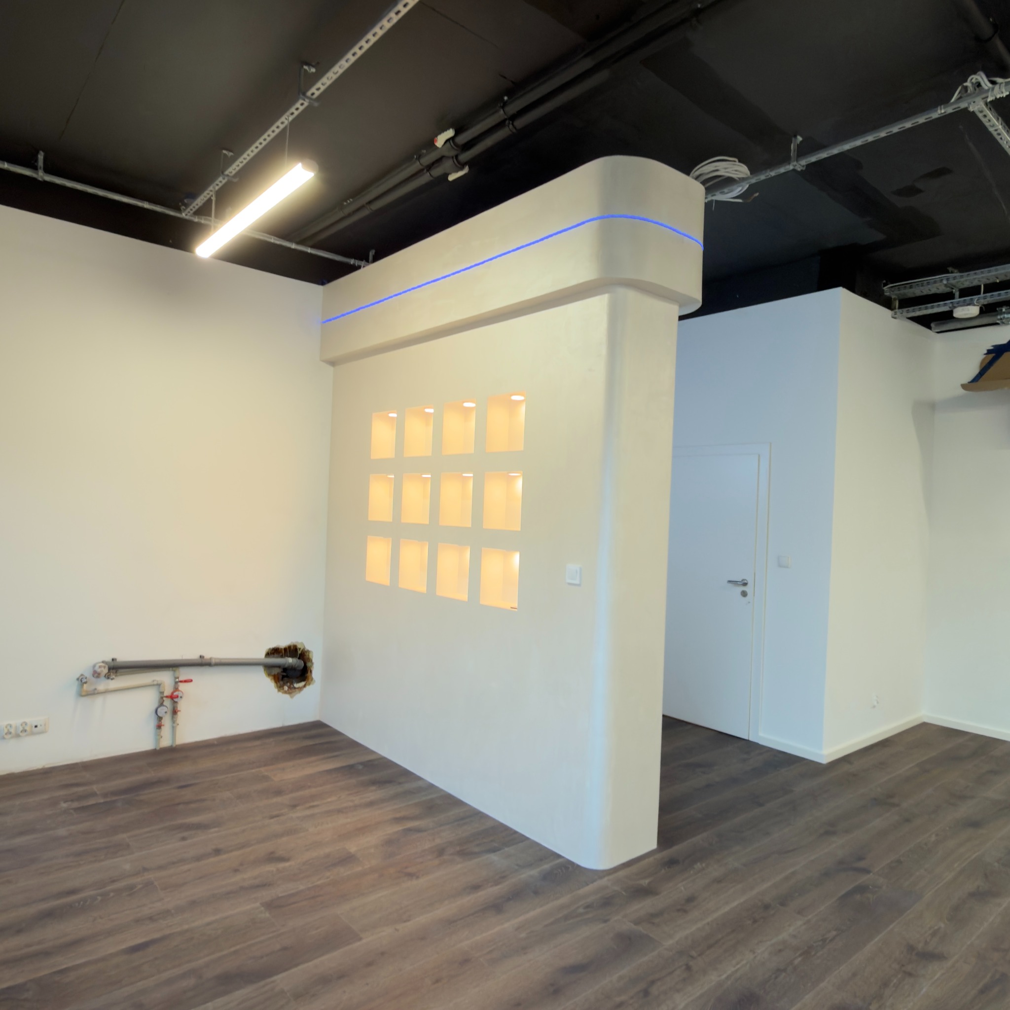 Nowoczesne wykończenie wnętrza biura: biała ściana działowa z podświetlanymi niszami, ciemna podłoga imitująca drewno, widoczne instalacje pod sufitem.