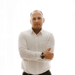 DOM UBEZPIECZENIOWY SPECTRUM AGENT W BRZOZOWIE - Portret mężczyzny w białej koszuli i czarnych spodniach, z zegarkiem na ręku, stojącego na tle jasnego okna. Ujęcie od pasa w górę, skrzyżowane ręce.
