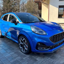 Glossper Auto Detailing - Błękitny Ford Puma ST na kostce brukowej przed domem. Widoczne detale: czarne felgi, czerwone zaciski hamulcowe, logo ST na grillu. Słoneczny dzień.