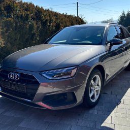 Glossper Auto Detailing - Szary samochód Audi A4 Avant zaparkowany na kostce brukowej w słoneczny dzień. Widoczny z przodu, z detalami reflektorów i logo. Tło stanowi zielony żywopłot i fragment domu.