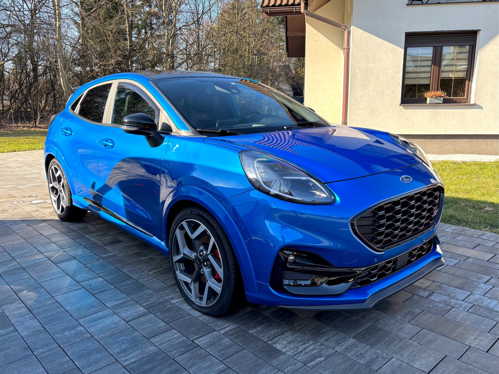 Błękitny Ford Puma ST na kostce brukowej przed domem. Widoczne detale: czarne felgi, czerwone zaciski hamulcowe, logo ST na grillu. Słoneczny dzień.