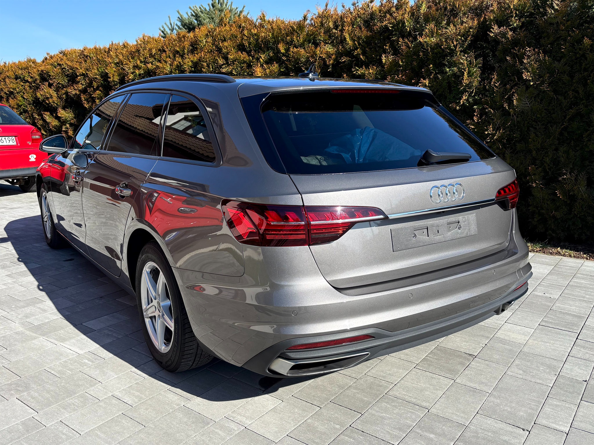 Szary samochód Audi A4 Avant, widok z tyłu na tle żywopłotu. Błyszcząca karoseria po umyciu, czyste felgi i opony. Auto zaparkowane na szarej kostce brukowej.