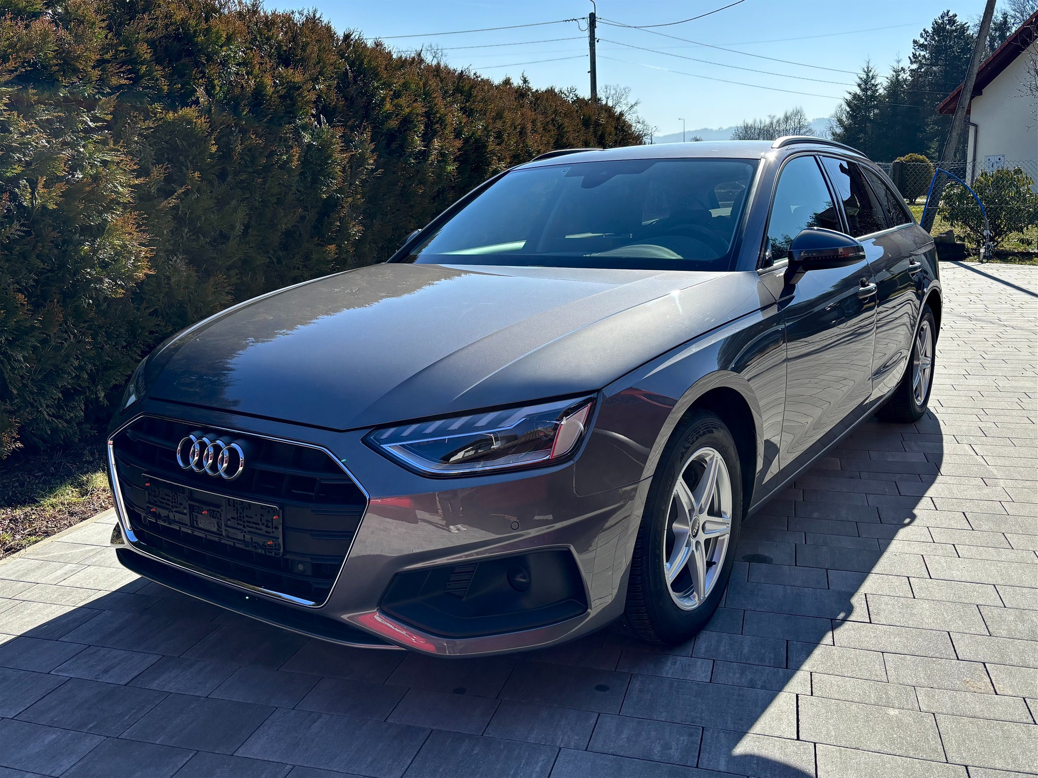 Szary samochód Audi A4 Avant zaparkowany na kostce brukowej w słoneczny dzień. Widoczny z przodu, z detalami reflektorów i logo. Tło stanowi zielony żywopłot i fragment domu.