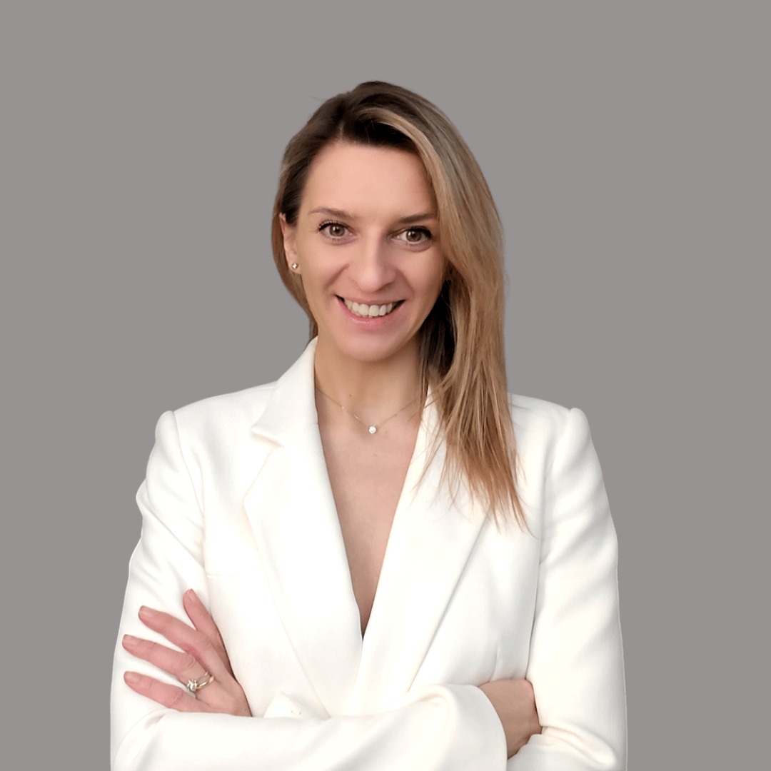 Portret uśmiechniętej kobiety w białym żakiecie, skrzyżowane ręce, subtelna biżuteria, jednolite szare tło. Profesjonalny wizerunek.