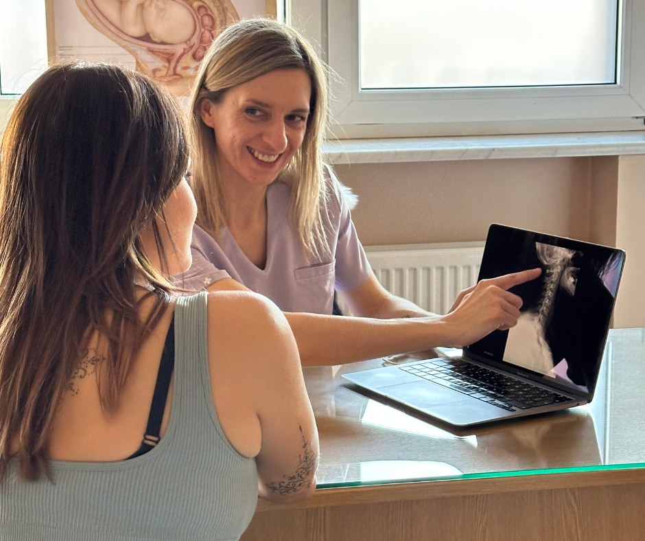 Konsultacja pacjentki: uśmiechnięta kobieta w fiolecie wskazuje na zdjęcie RTG kręgosłupa szyjnego na laptopie, siedząc przy biurku z pacjentką.