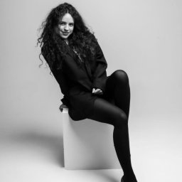 YOUR CONCEPT SPÓŁKA Z OGRANICZONĄ ODPOWIEDZIALNOŚCIĄ - Czarno-białe zdjęcie kobiety z kręconymi włosami, siedzącej na białym sześcianie w czarnym żakiecie i rajstopach, z delikatnym uśmiechem i spojrzeniem w obiektyw.