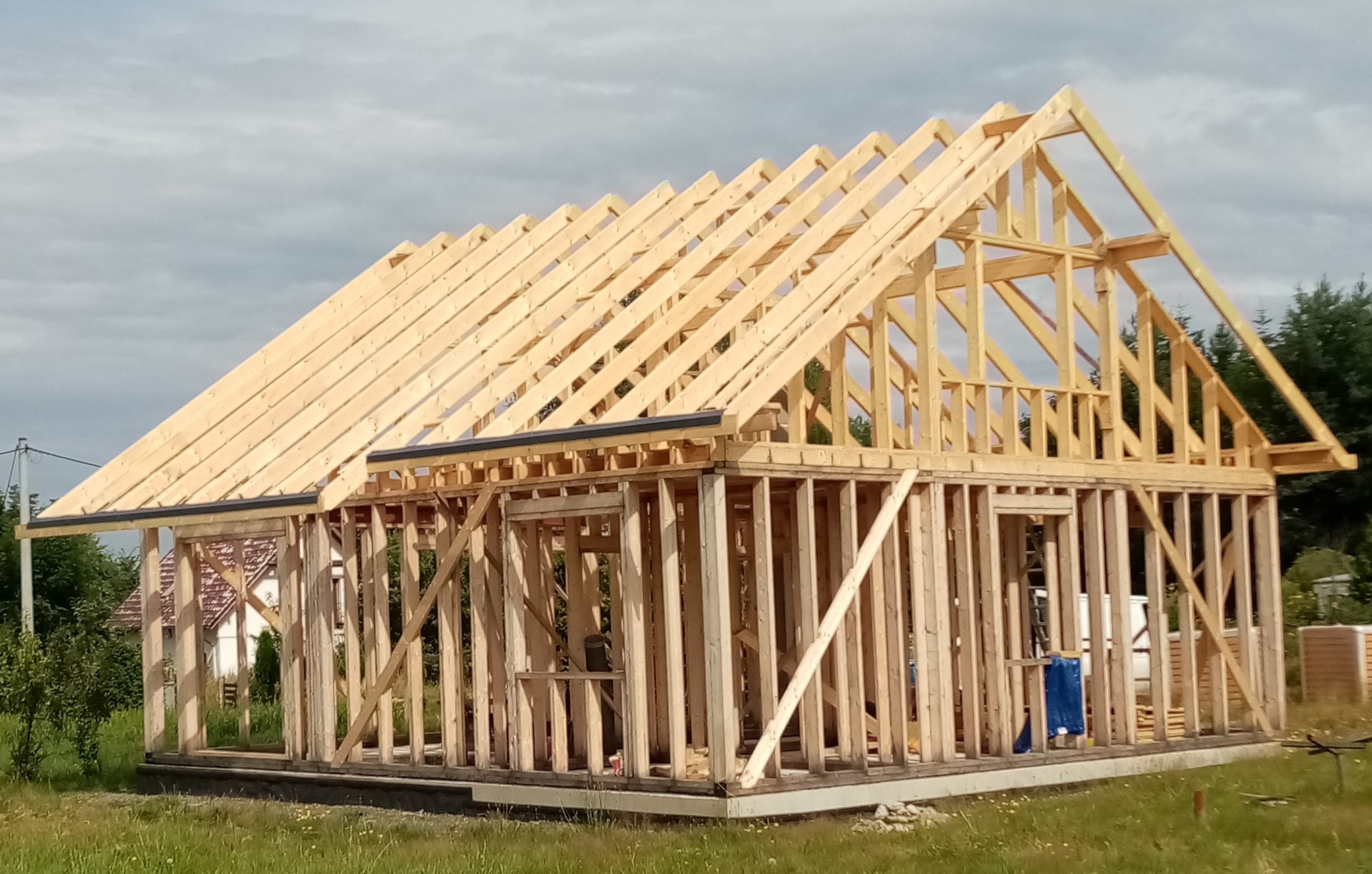 Konstrukcja szkieletowa domu w trakcie budowy, widoczne drewniane elementy ścian i dachu na tle zieleni i pochmurnego nieba. Etap montażu więźby dachowej.