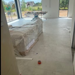 Krzysztof Kuśnierz - Jasne płytki podłogowe w nowym domu z dużymi oknami. Częściowo rozpakowane materiały budowlane. Widoczny fragment listwy przypodłogowej i taśma malarska.