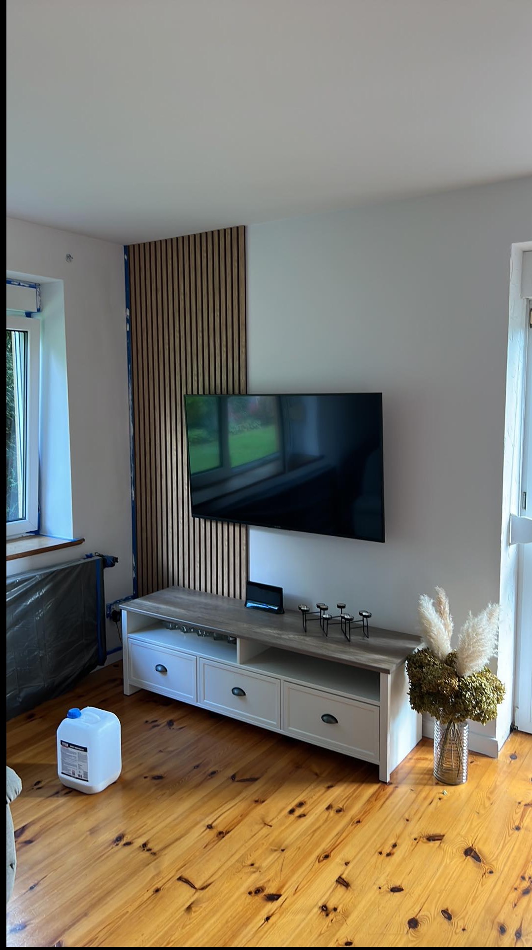 Salon po odświeżeniu: drewniana podłoga, białe ściany, lamelowa ściana TV, biała komoda. Widoczny pojemnik z farbą, sugerujący prace wykończeniowe.