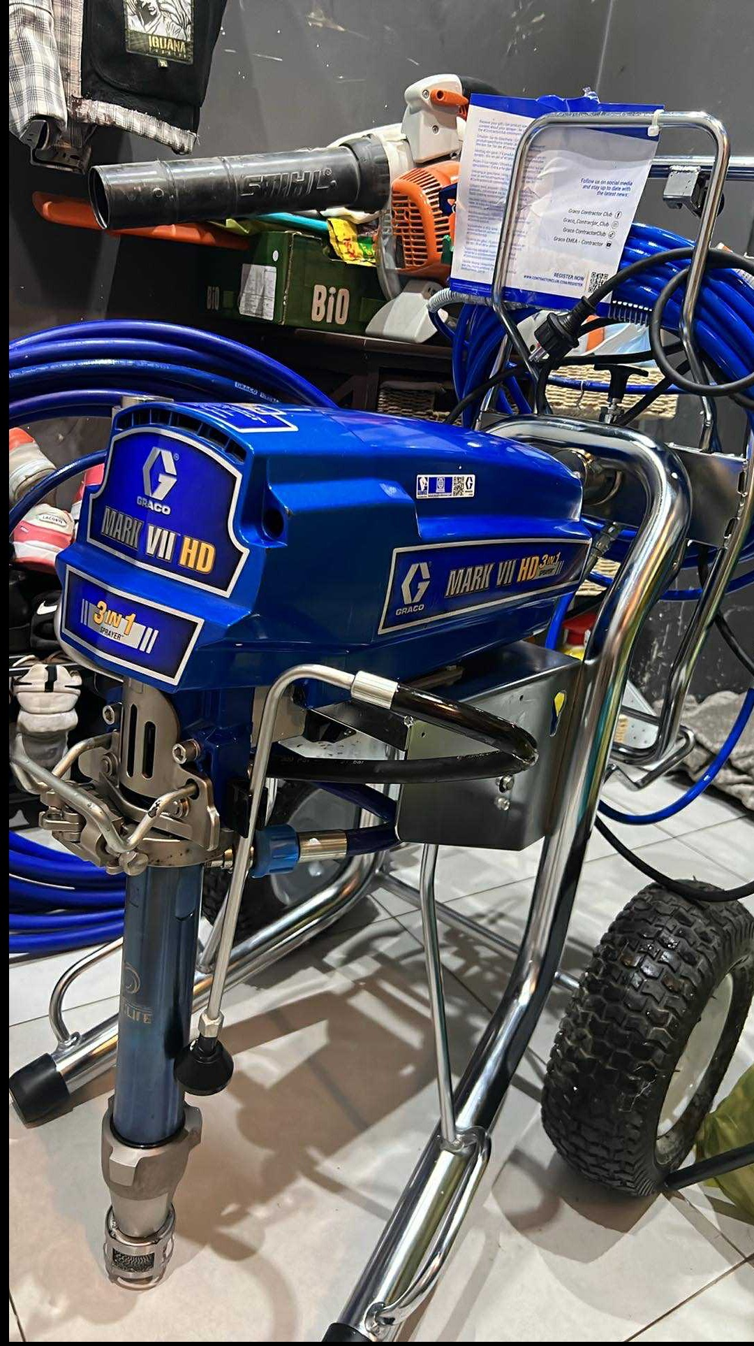 Profesjonalny agregat malarski Graco Mark VII HD na wózku z kołami, z niebieskim korpusem i chromowanymi elementami, otoczony przewodami. W tle widoczne narzędzia.