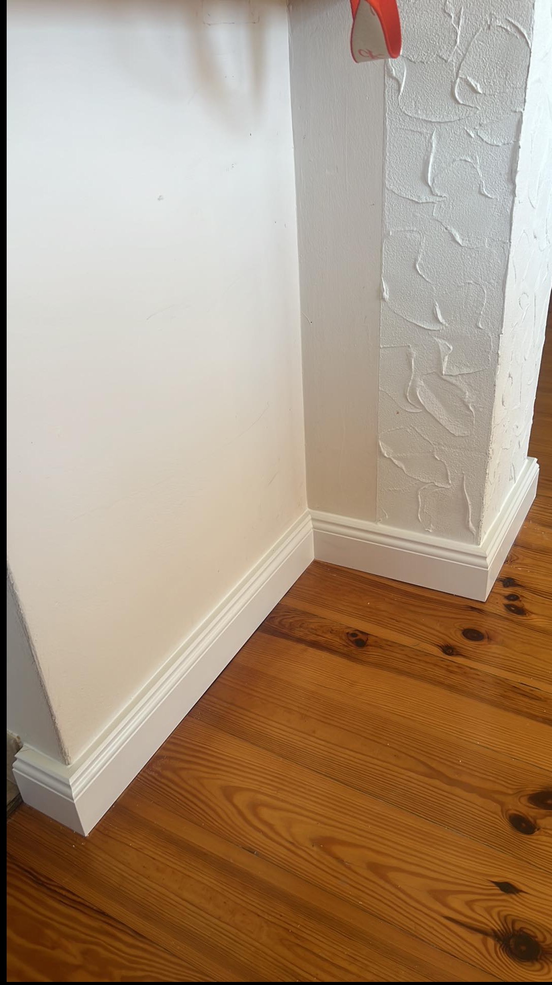 Białe listwy przypodłogowe przy ścianie z gładzią strukturalną i drewnianej podłodze. Detal wykończenia wnętrza, widok z dołu, ciepłe oświetlenie.