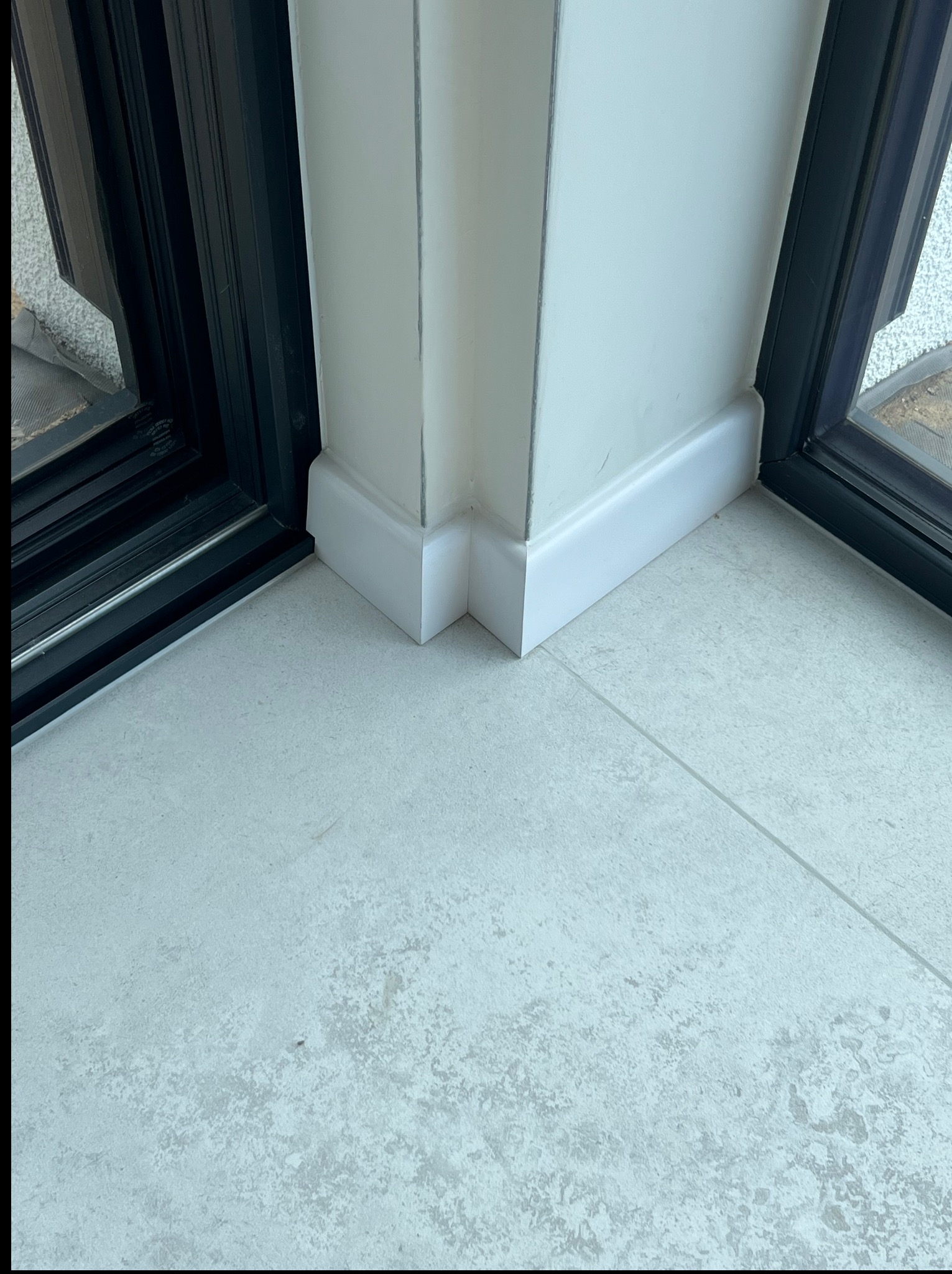 Białe listwy przypodłogowe narożne, łączące się z szarą, matową podłogą z płytek. Widoczne czarne ramy okienne. Minimalistyczny design.