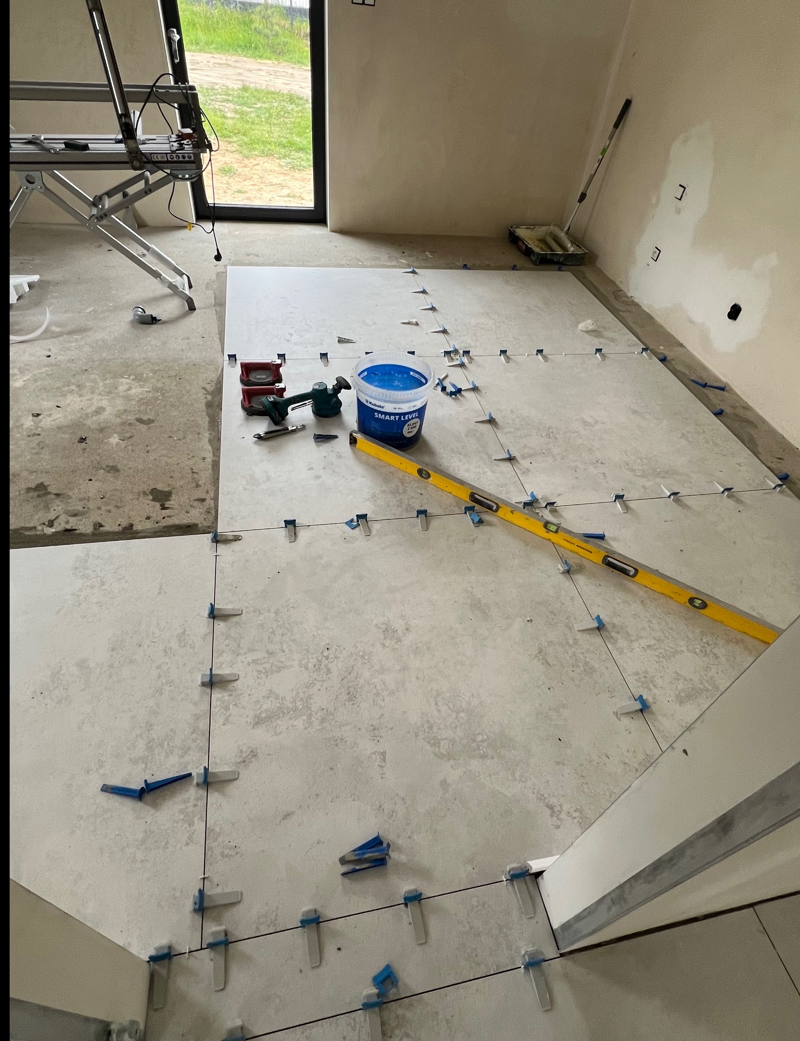 Układanie jasnych, kwadratowych płytek podłogowych z użyciem poziomicy i systemu poziomowania. Widoczne narzędzia glazurnicze i wiadro z klejem. Surowe ściany w tle.