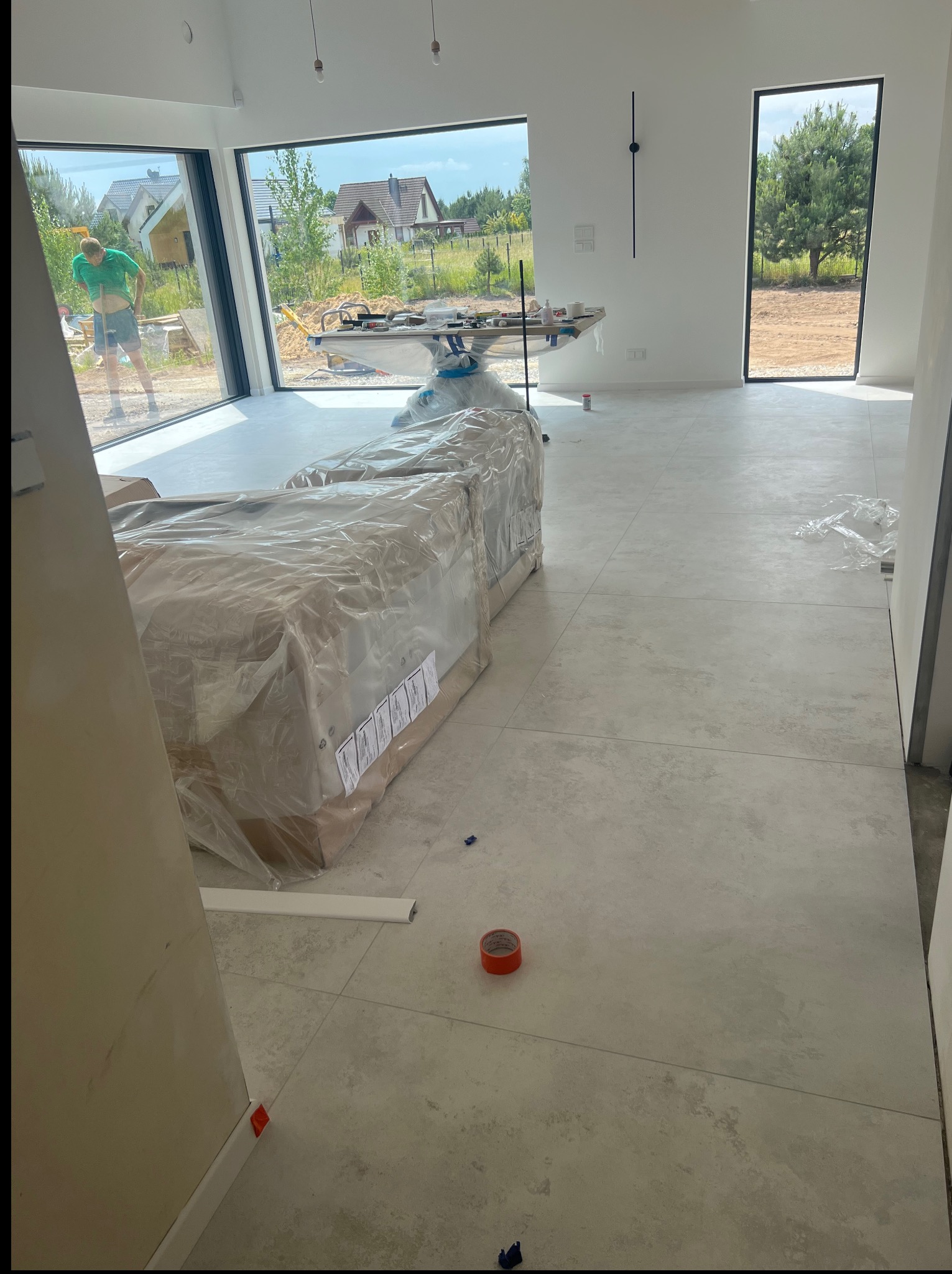 Jasne płytki podłogowe w nowym domu z dużymi oknami. Częściowo rozpakowane materiały budowlane. Widoczny fragment listwy przypodłogowej i taśma malarska.