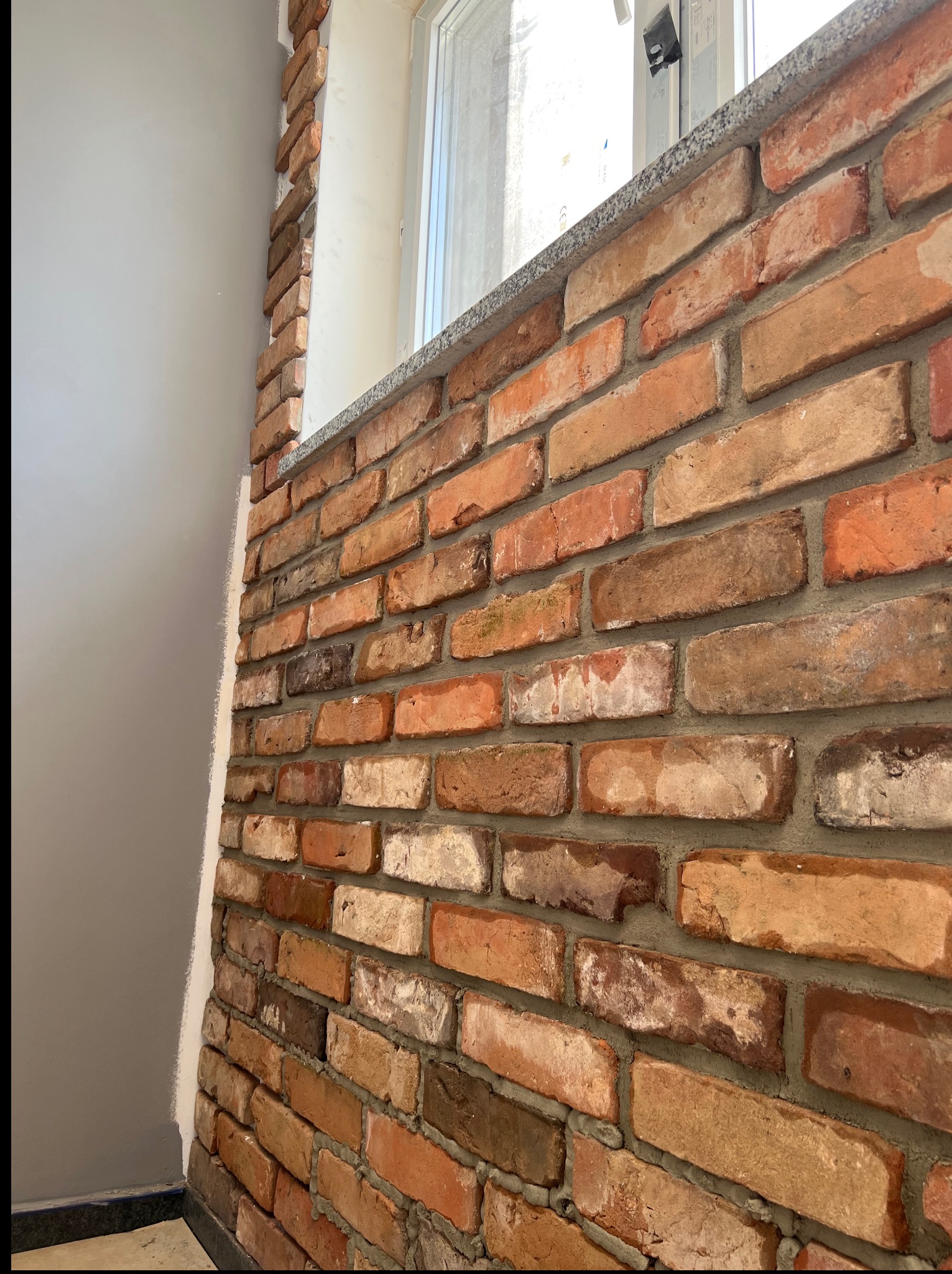 Ściana z cegły klinkierowej w odcieniach czerwieni i brązu, częściowo ułożona wokół okna. Widoczny fragment szarej ściany obok. Charakterystyczna faktura cegieł.