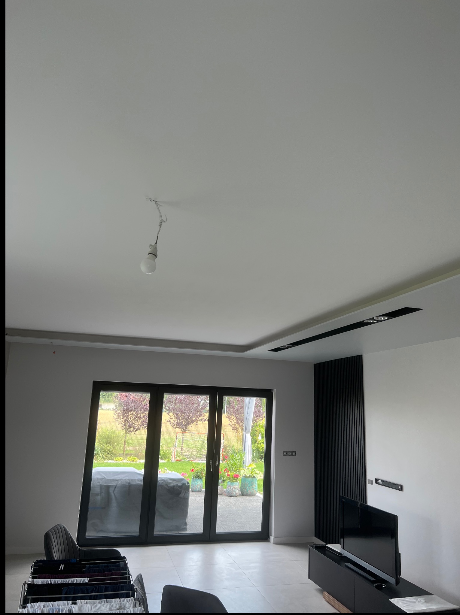 Wnętrze pokoju po remoncie: białe ściany, podwieszany sufit z oświetleniem, czarny panel dekoracyjny, duże okno z widokiem na ogród. W tle suszarka z praniem.