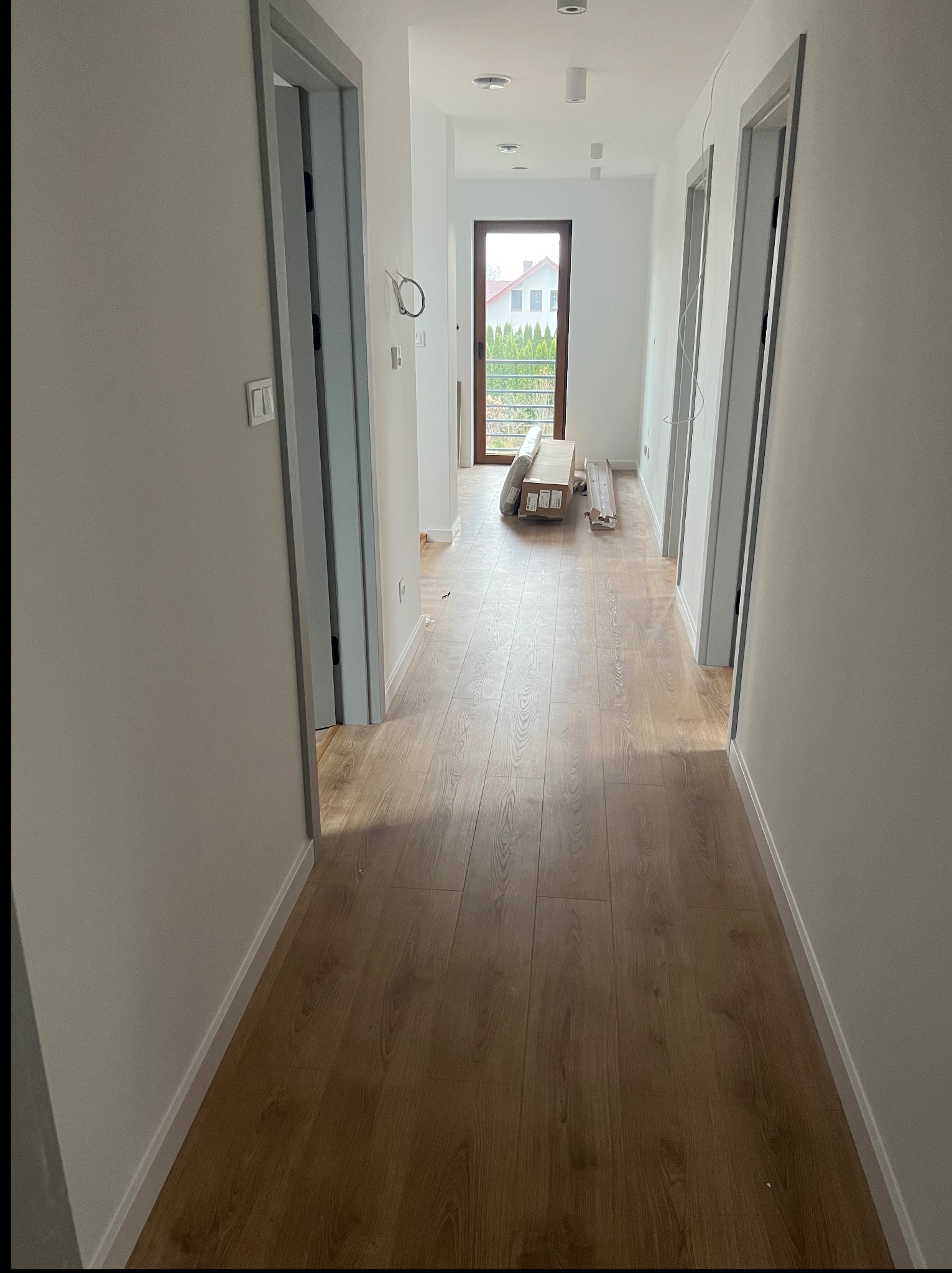 Korytarz po remoncie z panelami podłogowymi i szarymi ościeżnicami. W głębi widoczne materiały budowlane i okno z widokiem na zieleń.