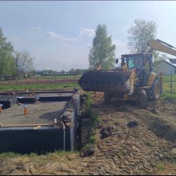 Green-Craft Ogrody - Żółta koparko-ładowarka zasypuje fundamenty budynku. Widoczne izolacje, rury kanalizacyjne i przygotowany teren pod budowę. W tle drzewa i zabudowania.