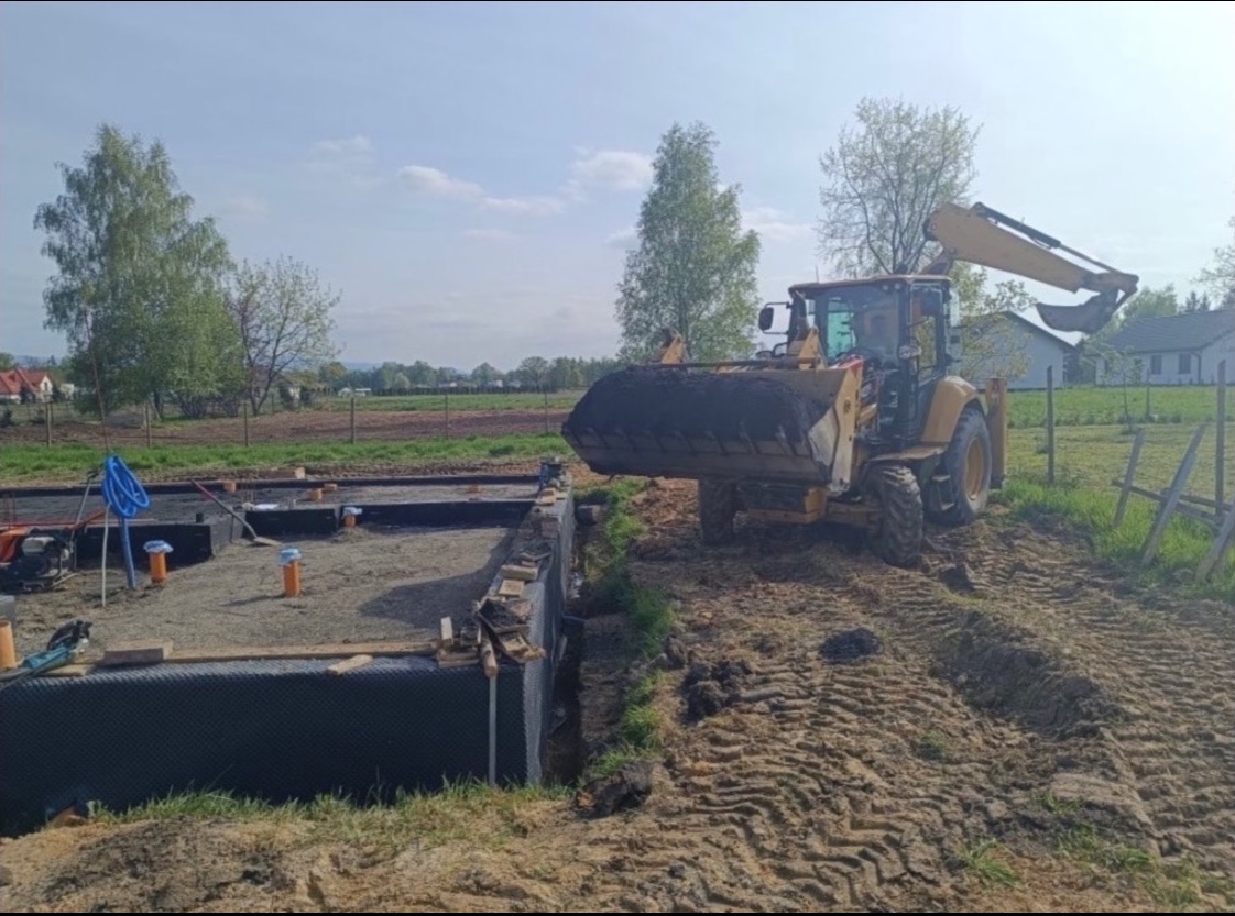 Żółta koparko-ładowarka zasypuje fundamenty budynku. Widoczne izolacje, rury kanalizacyjne i przygotowany teren pod budowę. W tle drzewa i zabudowania.
