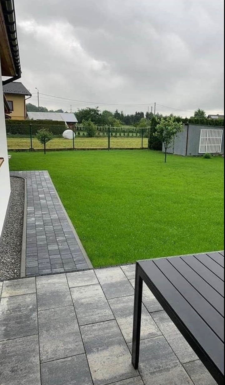 Szary chodnik z kostki brukowej prowadzi do zielonego trawnika. W tle widoczny dom i ogrodzenie. Na pierwszym planie fragment stołu ogrodowego.