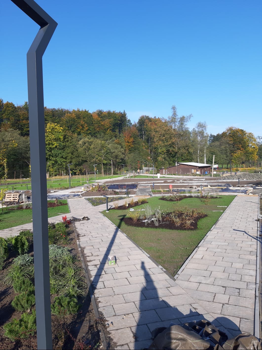 Park z alejkami z kostki, rabatami kwiatowymi i drzewami w tle, oświetlony słońcem. Widoczny element konstrukcyjny w lewym rogu kadru i cień osoby.