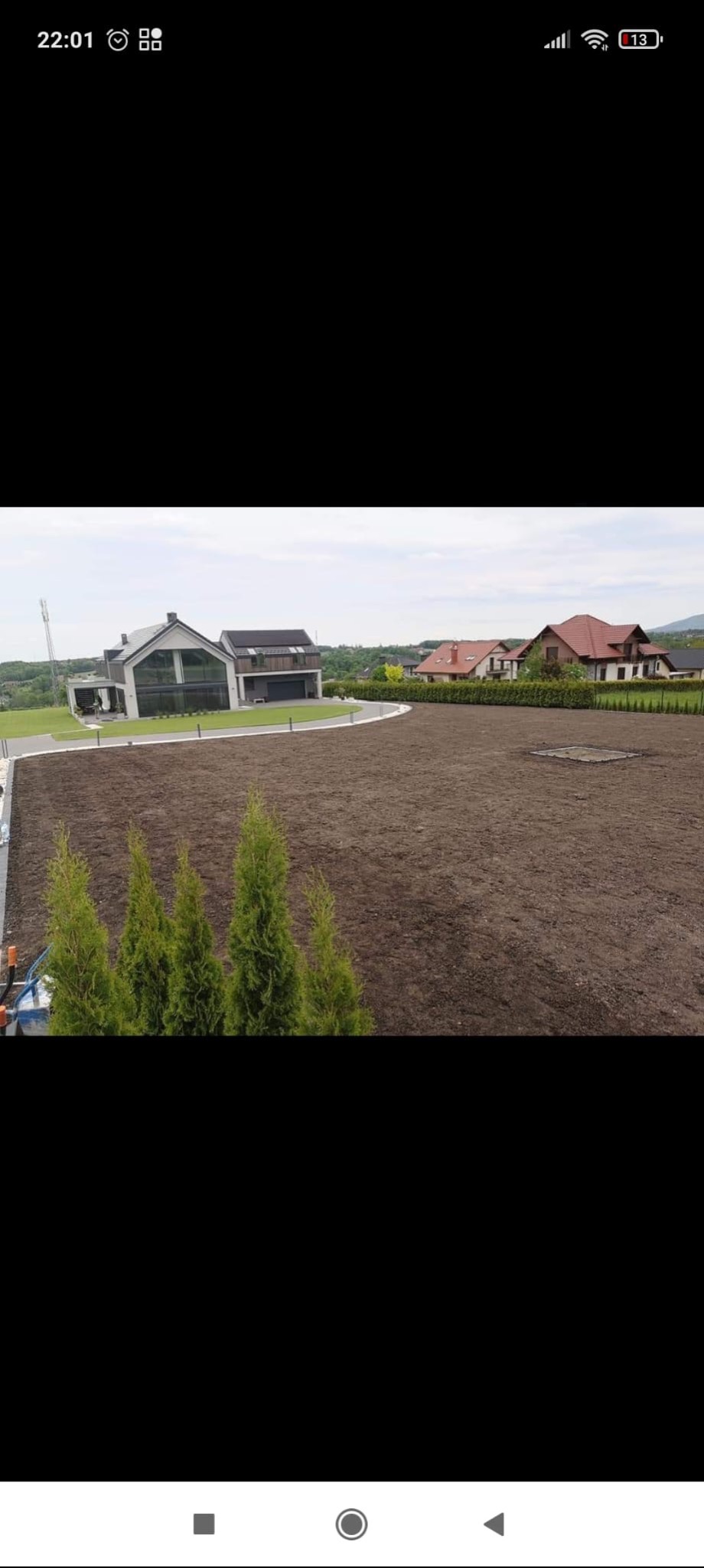 Przygotowany teren pod trawnik z posadzonymi tujami na pierwszym planie. W tle dom z nowoczesną architekturą i wybrukowany podjazd.