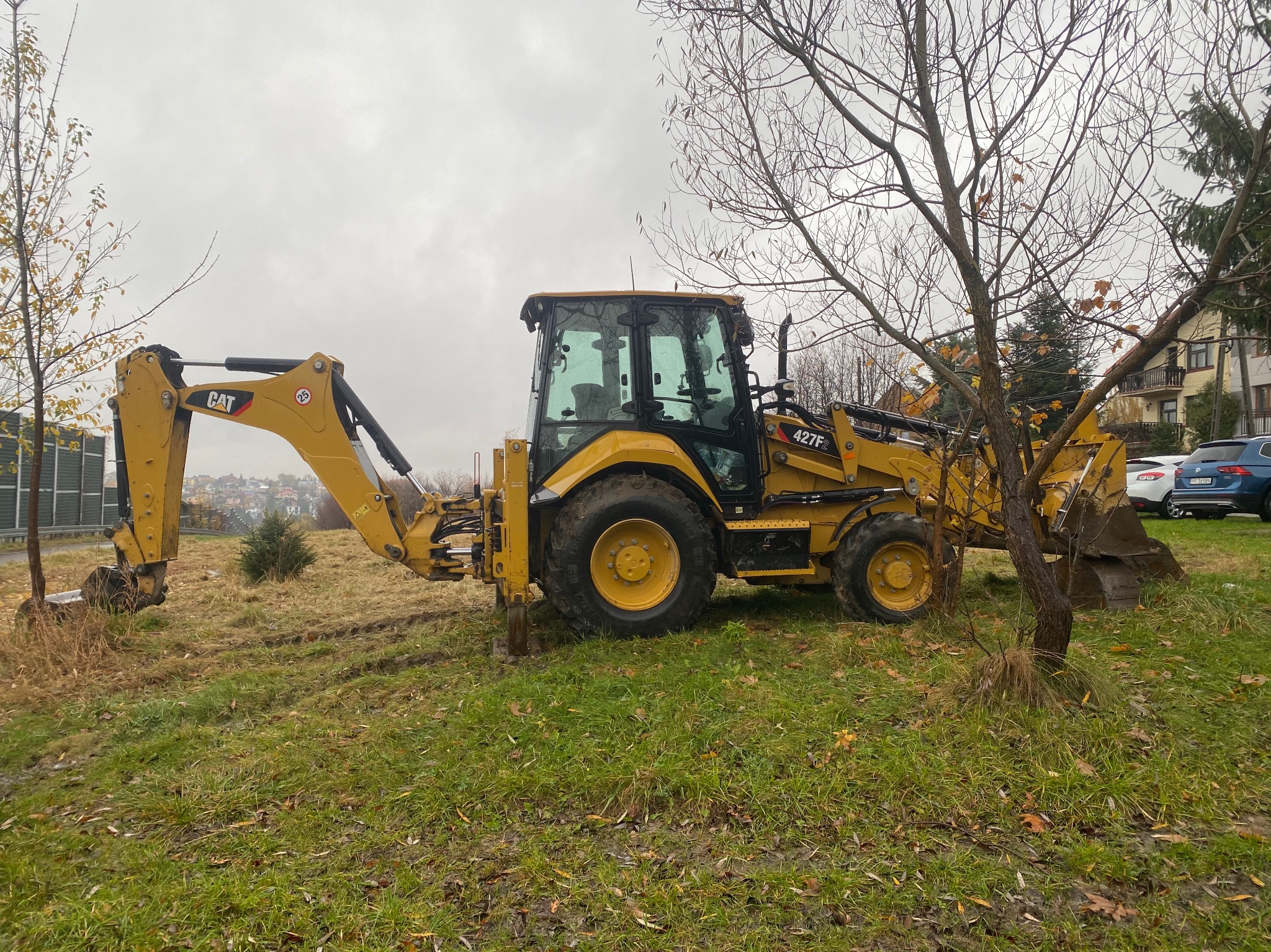 Żółta koparko-ładowarka CAT 427F2 na trawiastym terenie z drzewami w tle, w pochmurny dzień. Widoczne ślady opon i łyżka koparki.