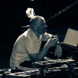 Tomasz Drozdowski - Czarno-białe zdjęcie muzyka z kubkiem, siedzącego przy pianinie i klawiszach Arturia, z nutami na pulpicie. Scena oświetlona, artystyczny kadr.