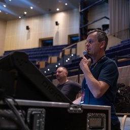 Tomasz Drozdowski - Mężczyzna z mikrofonem na tle widowni i sprzętu nagłośnieniowego. Ujęcie z dołu, widoczny fragment sceny i oświetlenie. Konferencja lub koncert.