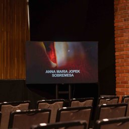 Tomasz Drozdowski - Sala koncertowa z rzędami krzeseł i ekranem wyświetlającym Annę Marię Jopek. Ceglana kolumna i drewniana ściana dodają charakteru wnętrzu. Scena gotowa na występ.