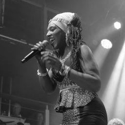 Tomasz Drozdowski FotoWideo - Czarno-białe zdjęcie piosenkarki z dredami, w bluzie w panterkę, śpiewającej do mikrofonu na scenie podczas koncertu, oświetlonej reflektorami.