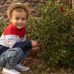 Tomasz Drozdowski FotoWideo - Uśmiechnięty chłopiec w kapeluszu kuca obok krzewu z czerwonymi owocami. Dziecko ma na sobie bluzę w kolorach czerwieni, bieli i granatu oraz dżinsy.