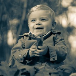 Tomasz Drozdowski FotoWideo - Uśmiechnięty blond chłopiec w kurtce siedzi w liściach, trzymając jeden w dłoniach. Zdjęcie w sepii, z rozmytym tłem drzew.