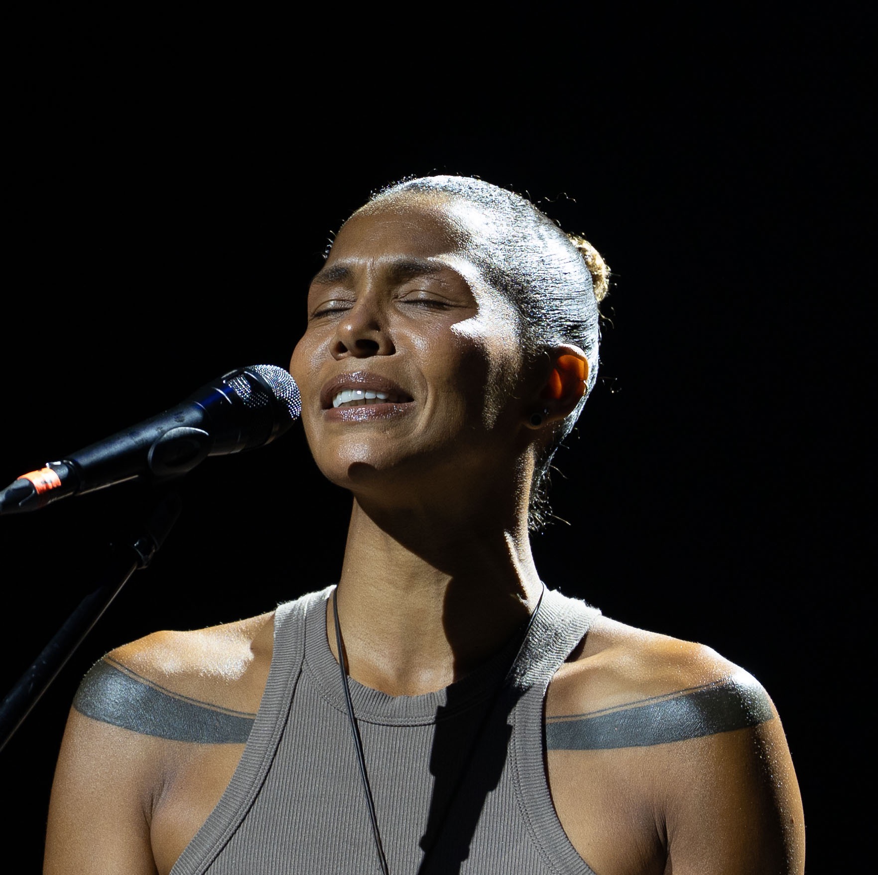 Portret kobiety z zamkniętymi oczami, śpiewającej do mikrofonu na scenie podczas koncertu. Widoczne tatuaże na ramionach i oświetlenie sceniczne.